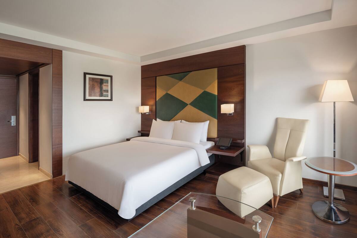 A hotel room with a large bed, a desk with a lamp, a chair, and a floor lamp.