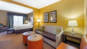 A hotel room with a bed, a sofa, two ottomans, a nightstand with a lamp, and two framed pictures on the wall. - Best Western Plus Tupelo Inn & Suites (Tupelo)
