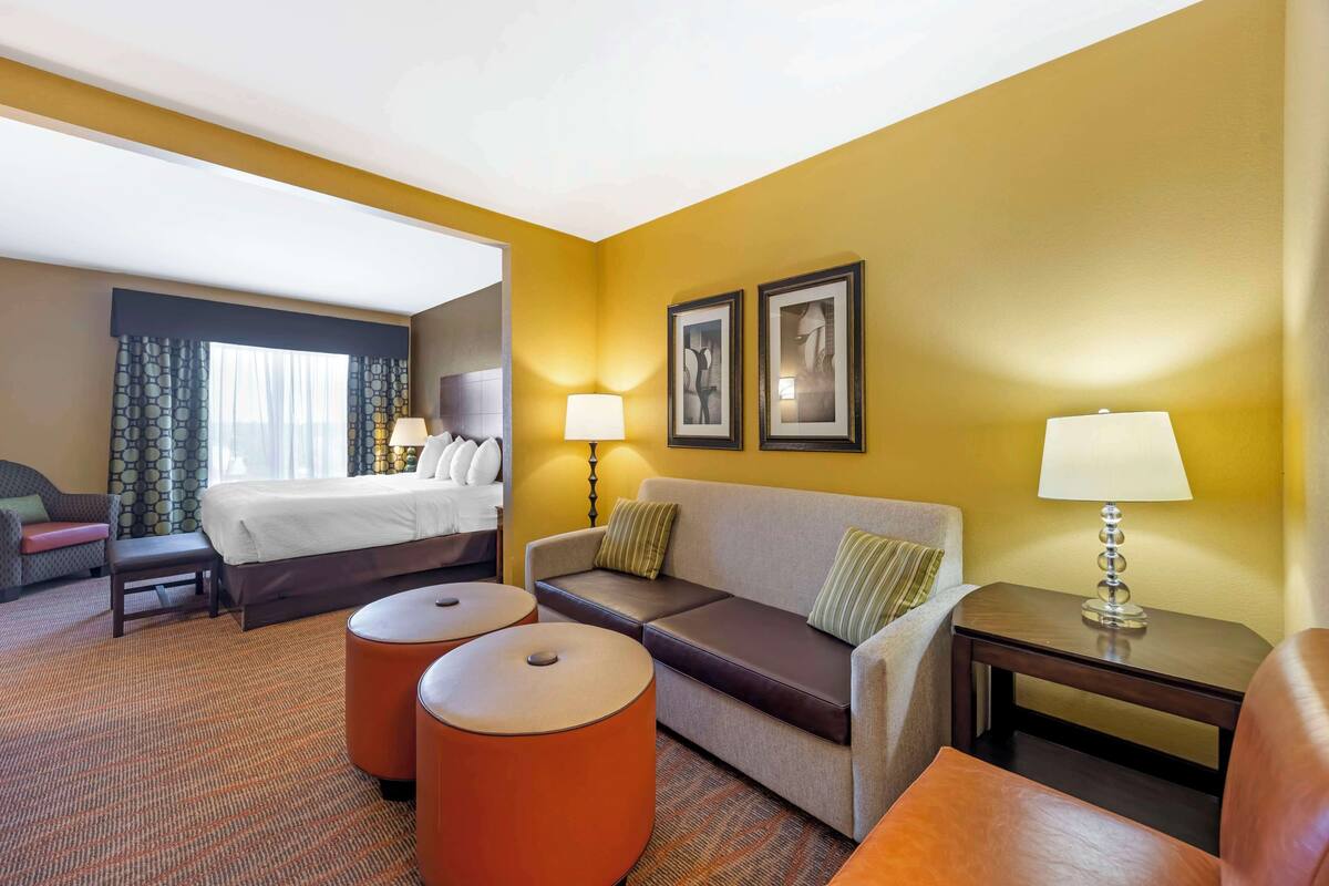 A hotel room with a bed, a sofa, two ottomans, a nightstand with a lamp, and two framed pictures on the wall.