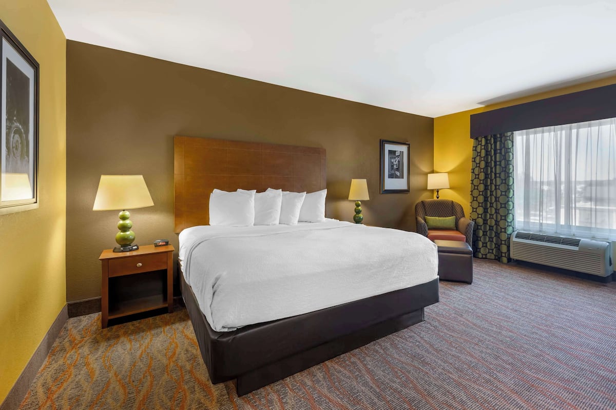 A hotel room with a large bed, two bedside tables with lamps, a chair, a window with curtains, and an air conditioning unit.