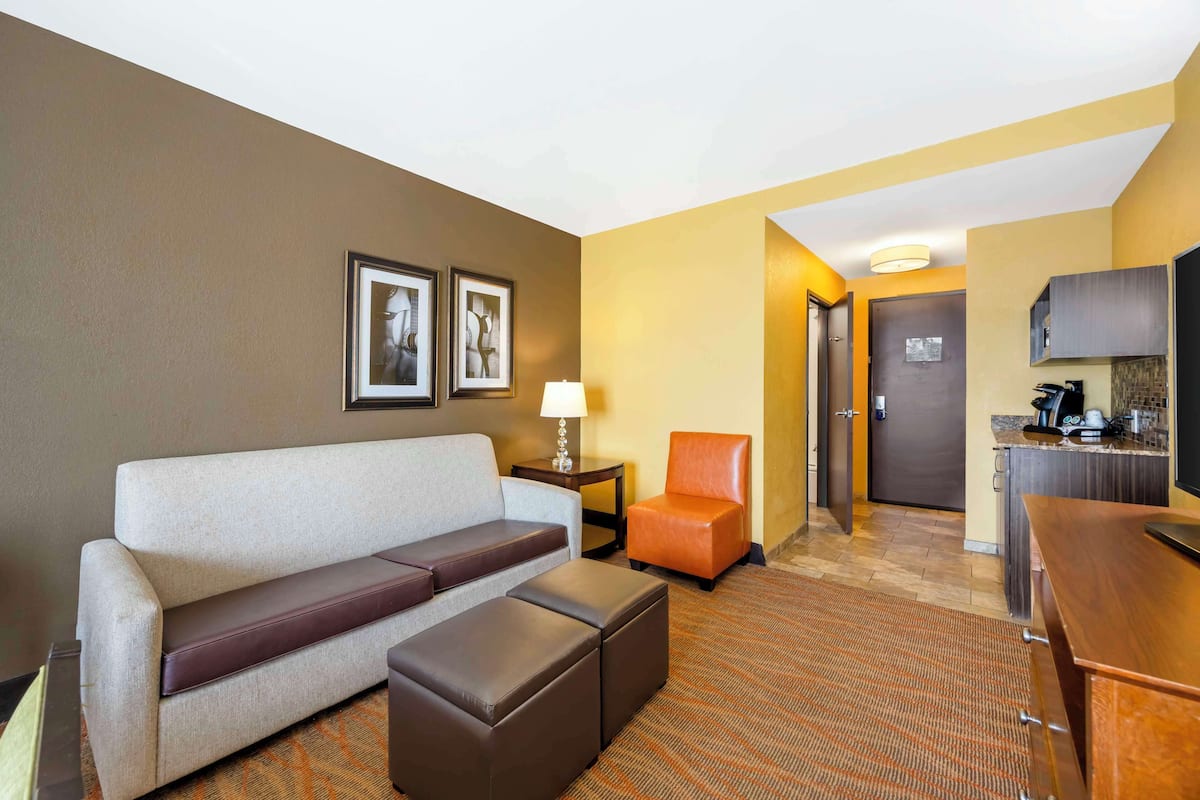 A hotel room with a sofa, ottoman, a small table with a lamp, a chair, and a kitchenette area.