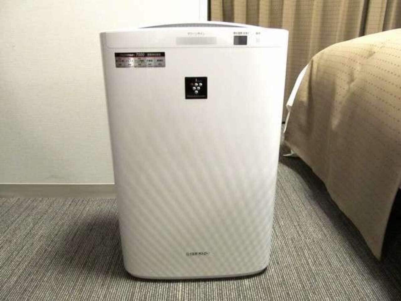 Calentador portátil blanco con panel de control y cable de alimentación, sobre un piso alfombrado en una habitación.