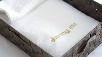 Toalla blanca con el nombre "dermy inn" bordado en dorado sobre un fondo blanco, colocada dentro de una cesta tejida.