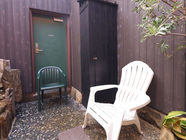 A small outdoor seating area with a green chair and a white chair, a door, and a stone wall.
