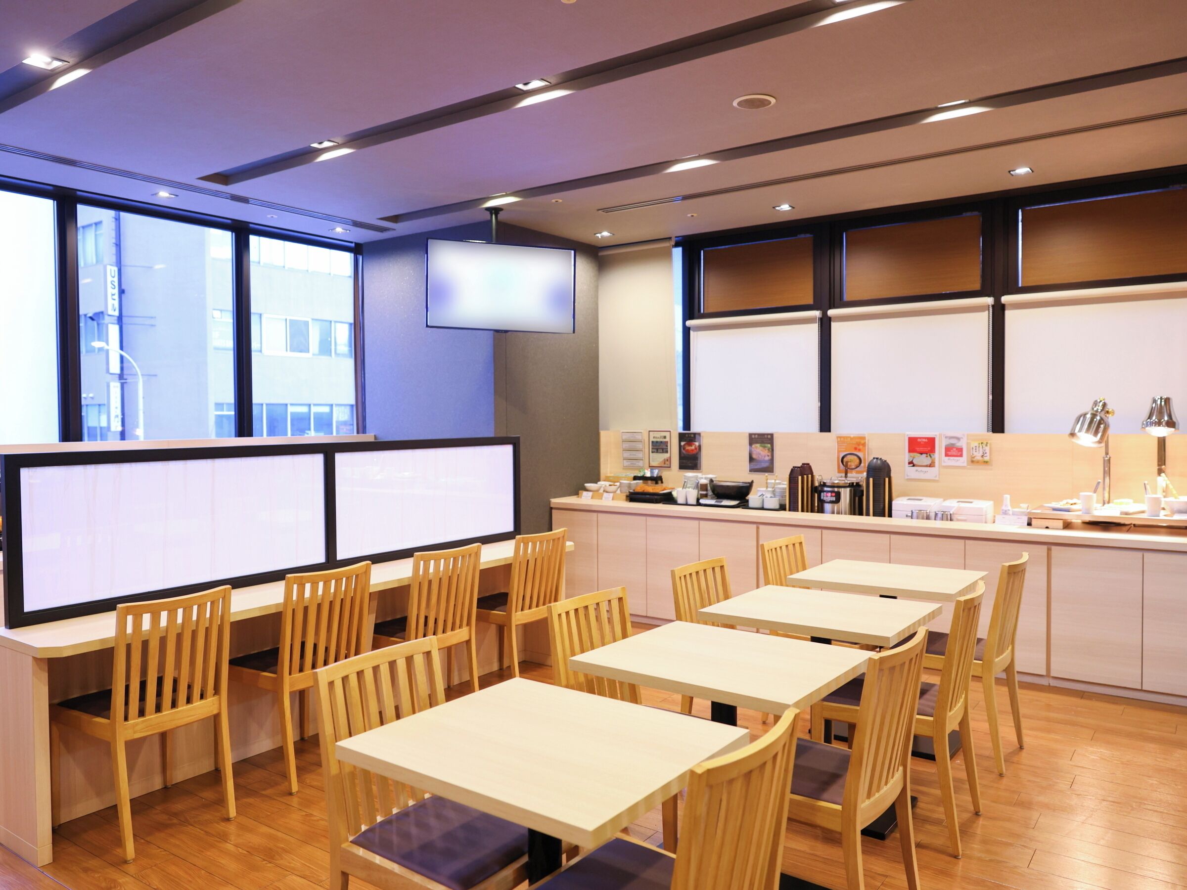 A modern dining area with wooden tables and chairs, a counter with coffee machines, and a large window with a view of the city.