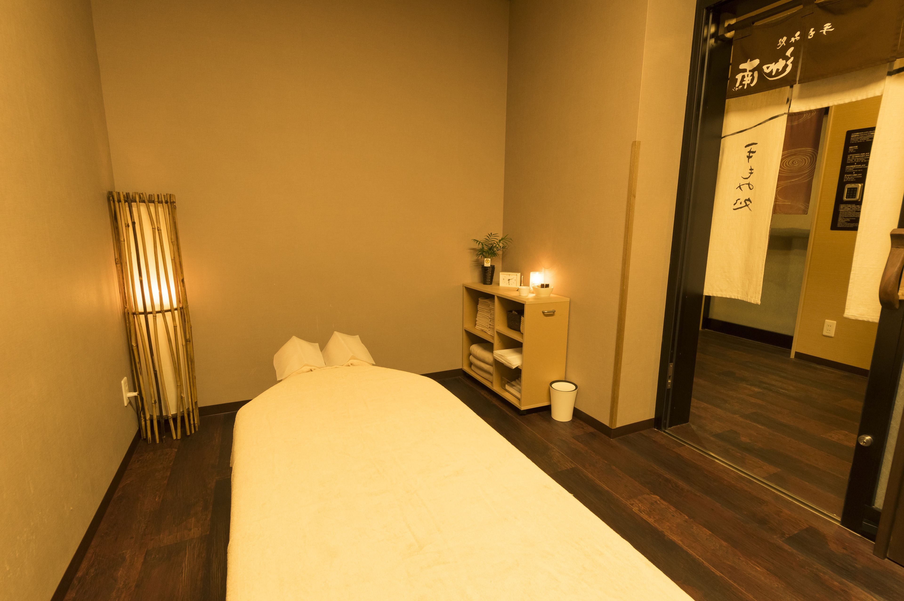 A hotel room with a bed, a bedside table with a lamp, a small shelf with towels, and a trash bin.