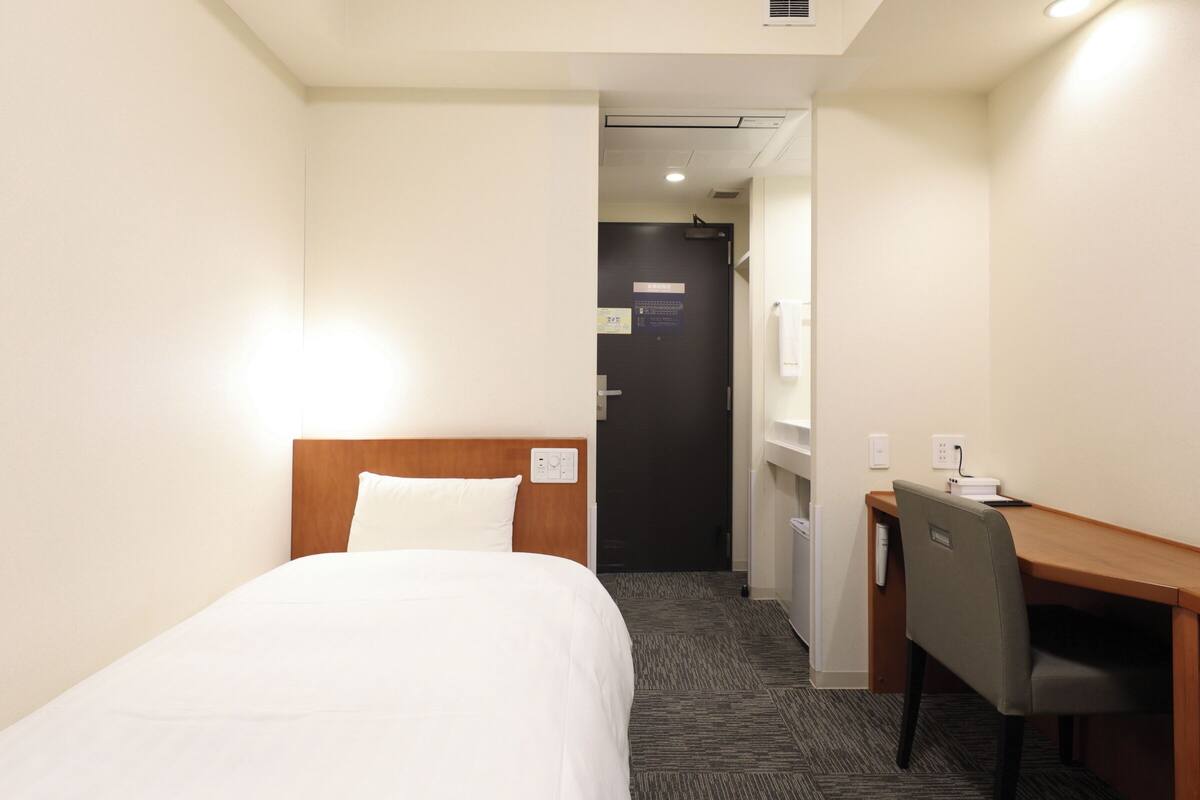 A hotel room with a bed, a desk, and a chair. There is a door leading to another room.