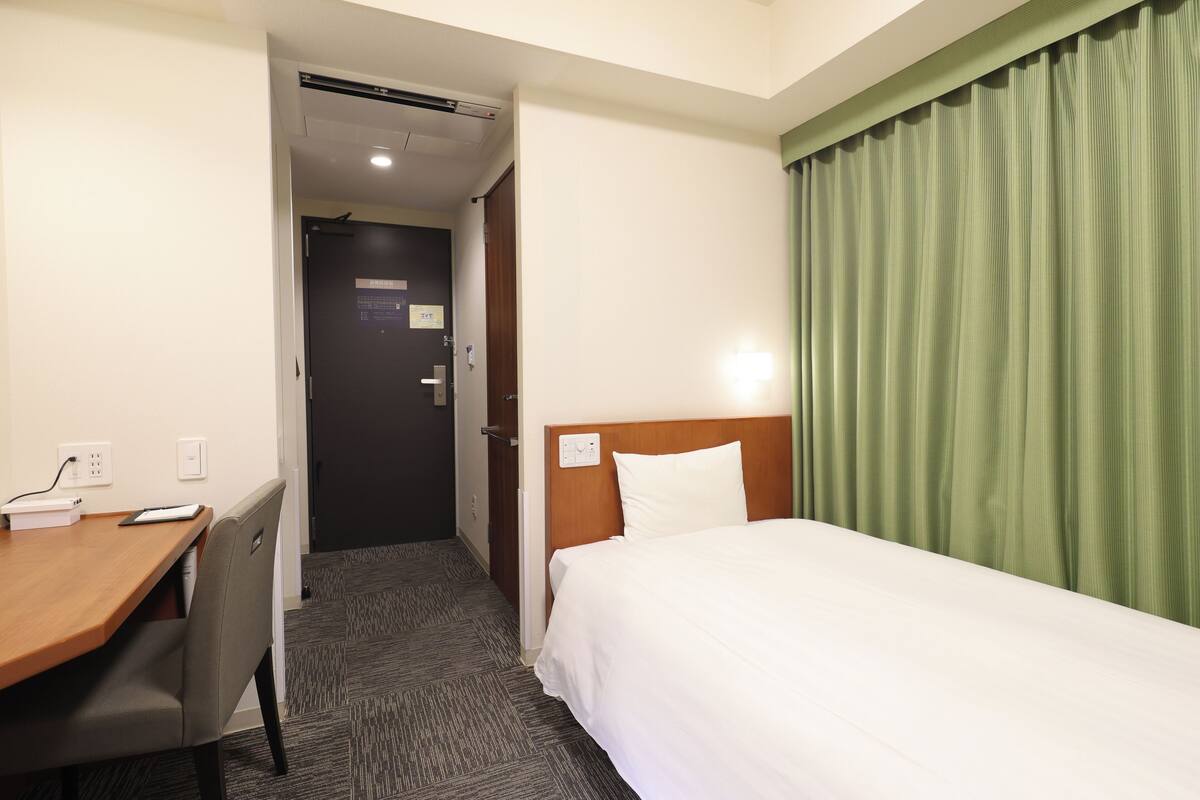 A hotel room with a wooden desk, a chair, a bed with a white headboard, and a green curtain.