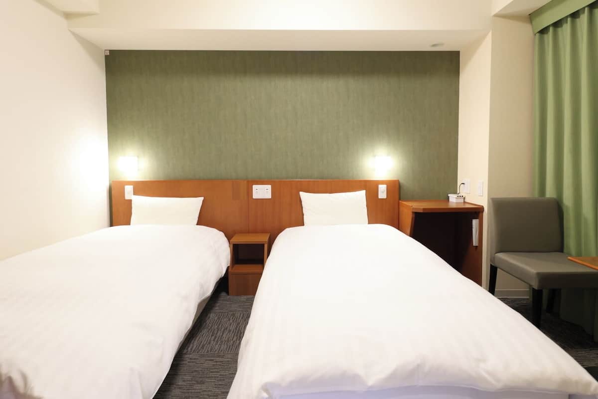 A hotel room with two single beds, a wooden headboard, a green curtain, a gray armchair, and a small table.