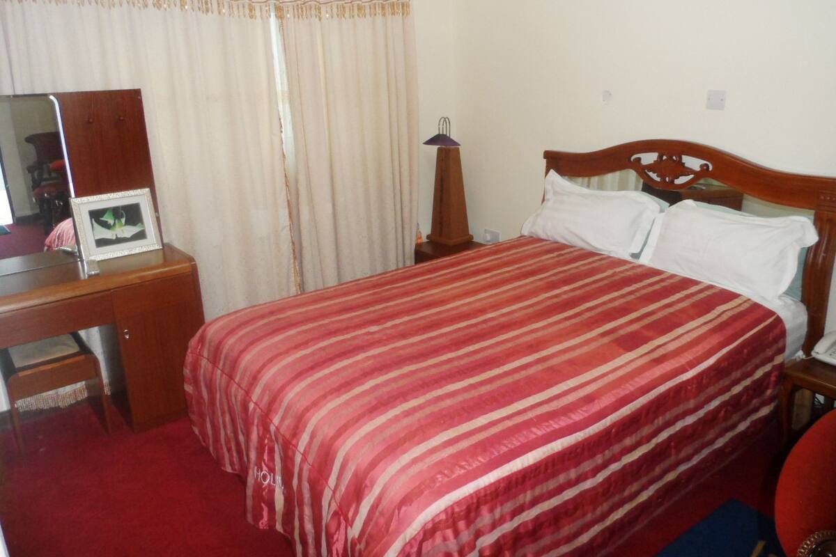 A bedroom with a bed, a wooden desk, a chair, a mirror, and a bedside table.