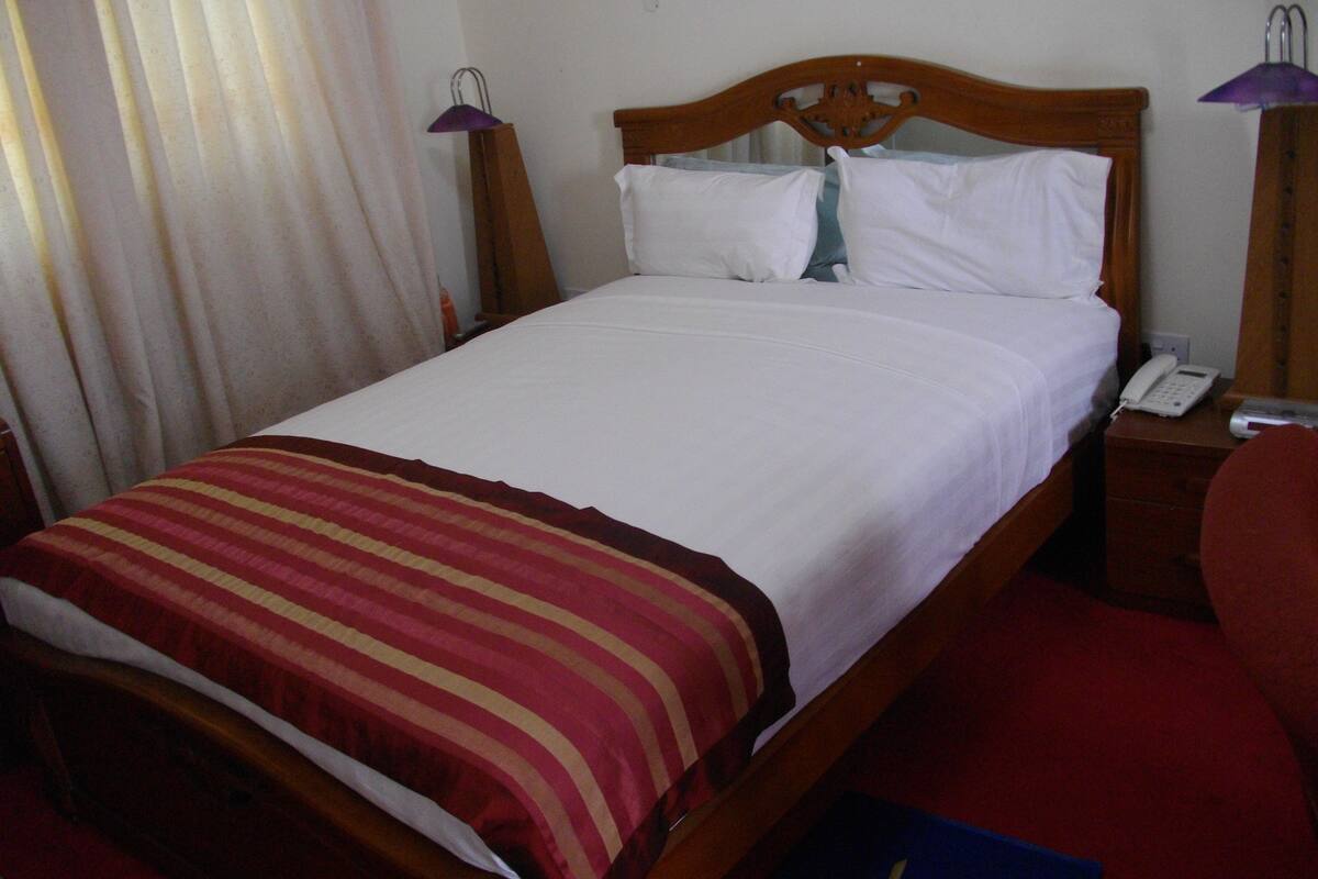 A standard hotel room with a wooden bed, white bedding, a striped throw blanket, a bedside table with a lamp, and a phone.