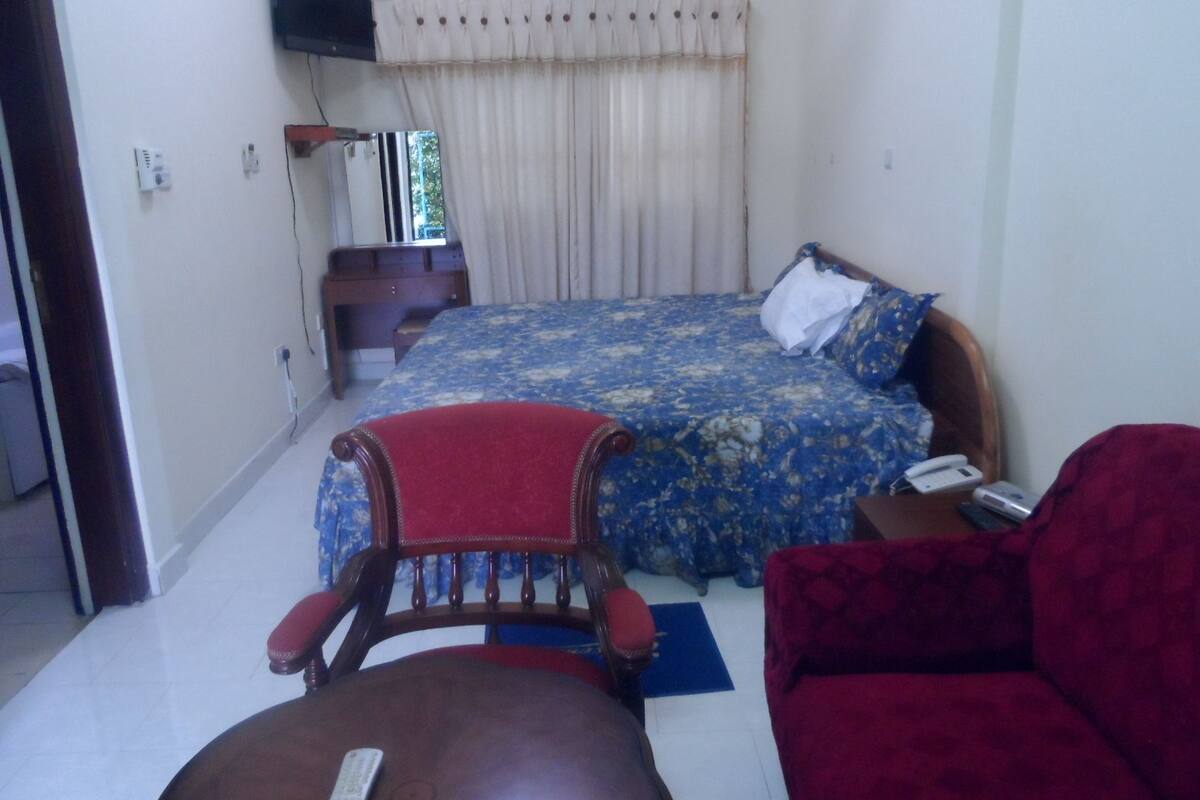A bedroom with a bed, a wooden chair, a red sofa, a nightstand, a TV, and an air conditioner.