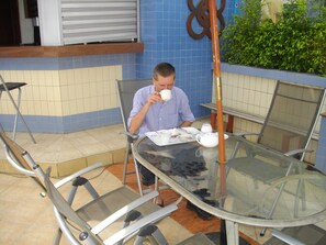 A man seated at a glass table with a chair, a white teapot, and a cup on the table. - De Holiday Beach Hotel (Kokrobite)