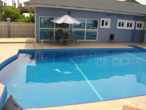 A swimming pool with a tiled deck, a lounge area with chairs and a striped umbrella, and a building with windows and a sign reading "OBAM TERRAC". - De Holiday Beach Hotel (Kokrobite)