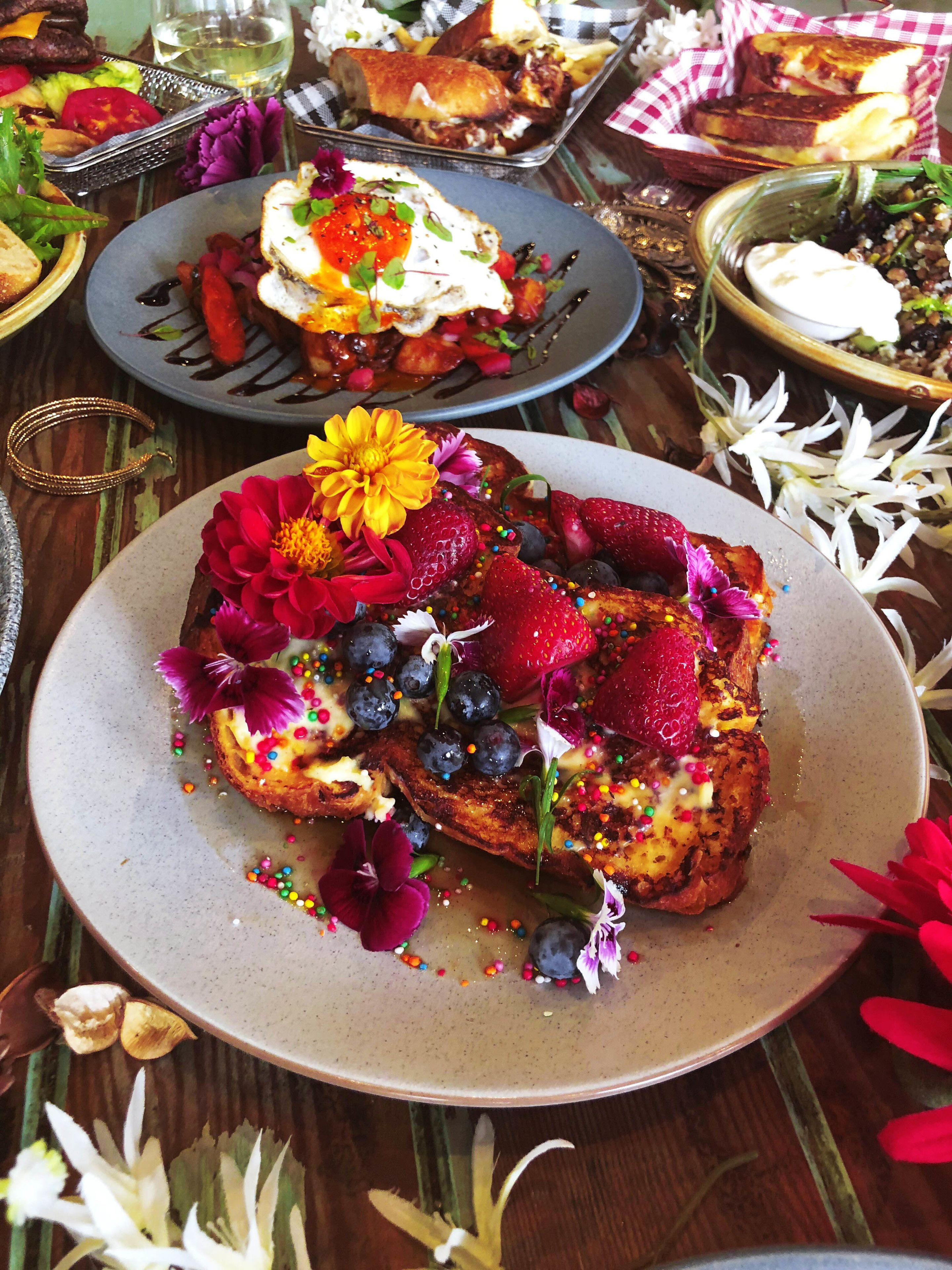 A breakfast setting with a plate of French toast topped with strawberries, blueberries, and a flower, accompanied by a side of pancakes and a glass of white wine.
