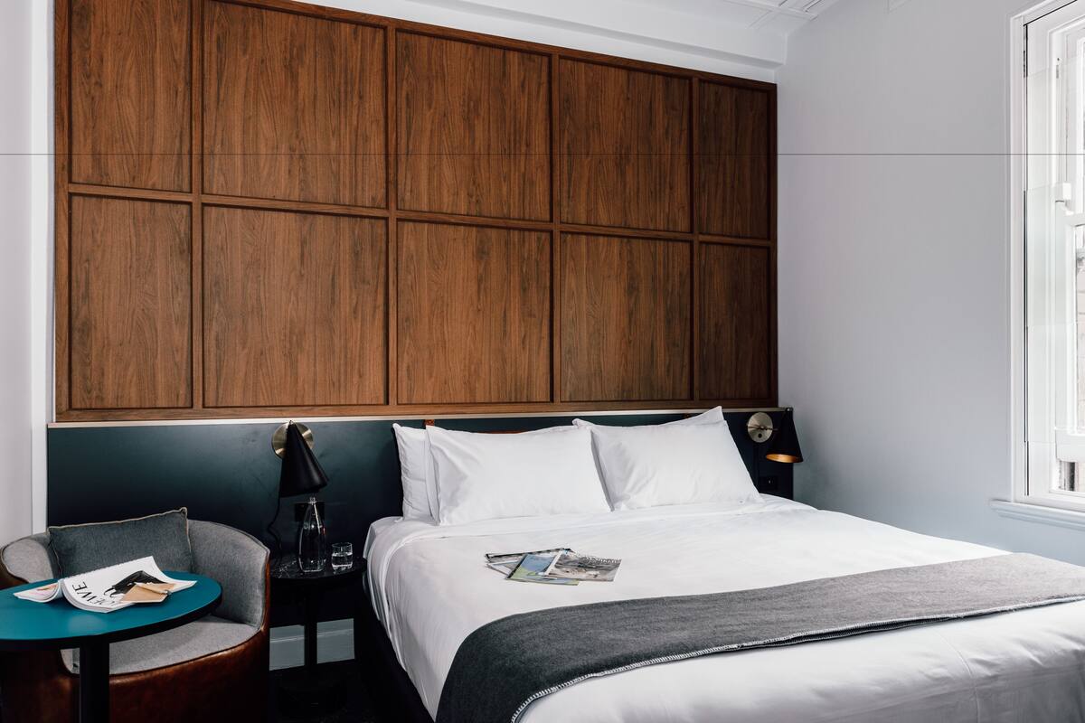 Une chambre d’hôtel avec un grand lit, une table de chevet avec une lampe, une petite table ronde avec une chaise, et une tête de lit en bois.
