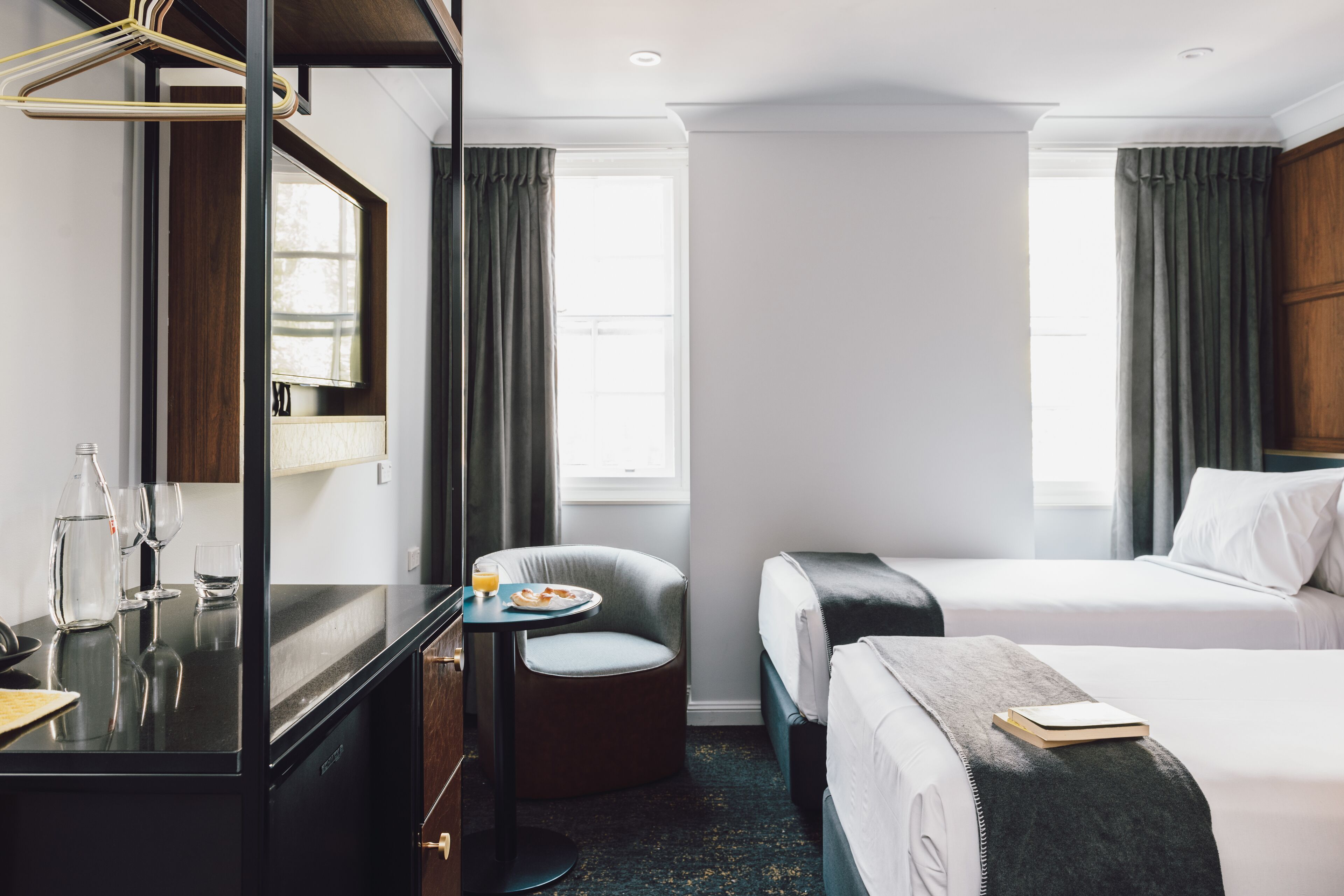 A hotel room with a bed, a desk with a chair, a small table with a plate of food, and a mirror.
