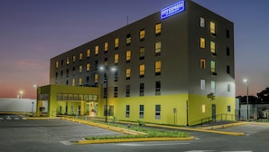 A multi-story hotel with a flat facade, a parking lot with marked spaces, and a sign indicating a 10-minute walk to the hotel. - City Express by Marriott Managua (Managua)