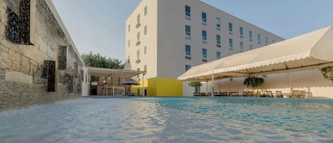Una piscina con zona techada, un edificio con múltiples ventanas y un muro de ladrillo.