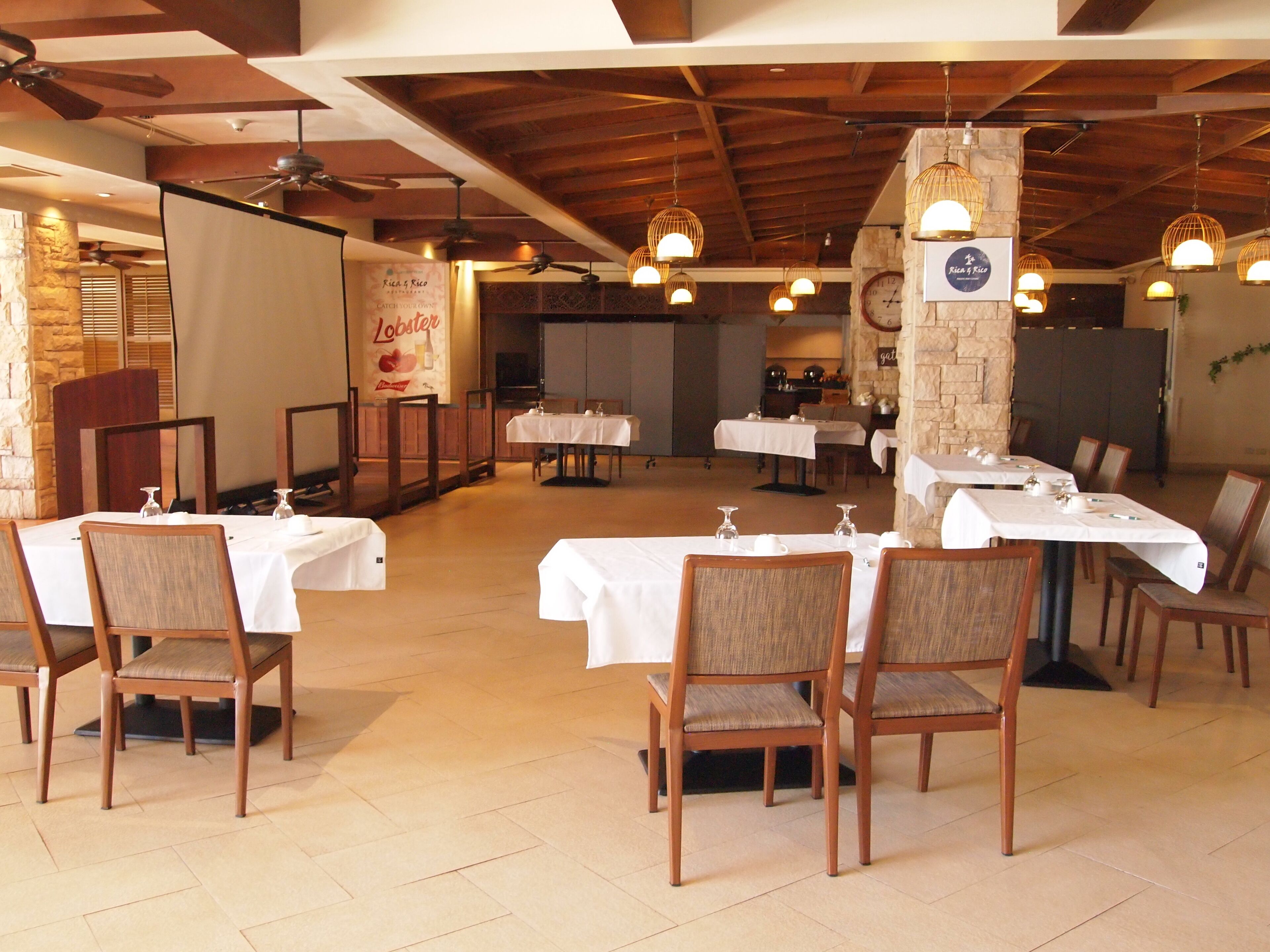 Un restaurant doté de nombreuses tables et chaises, de poutres en bois au plafond, de murs en pierre et de luminaires suspendus.