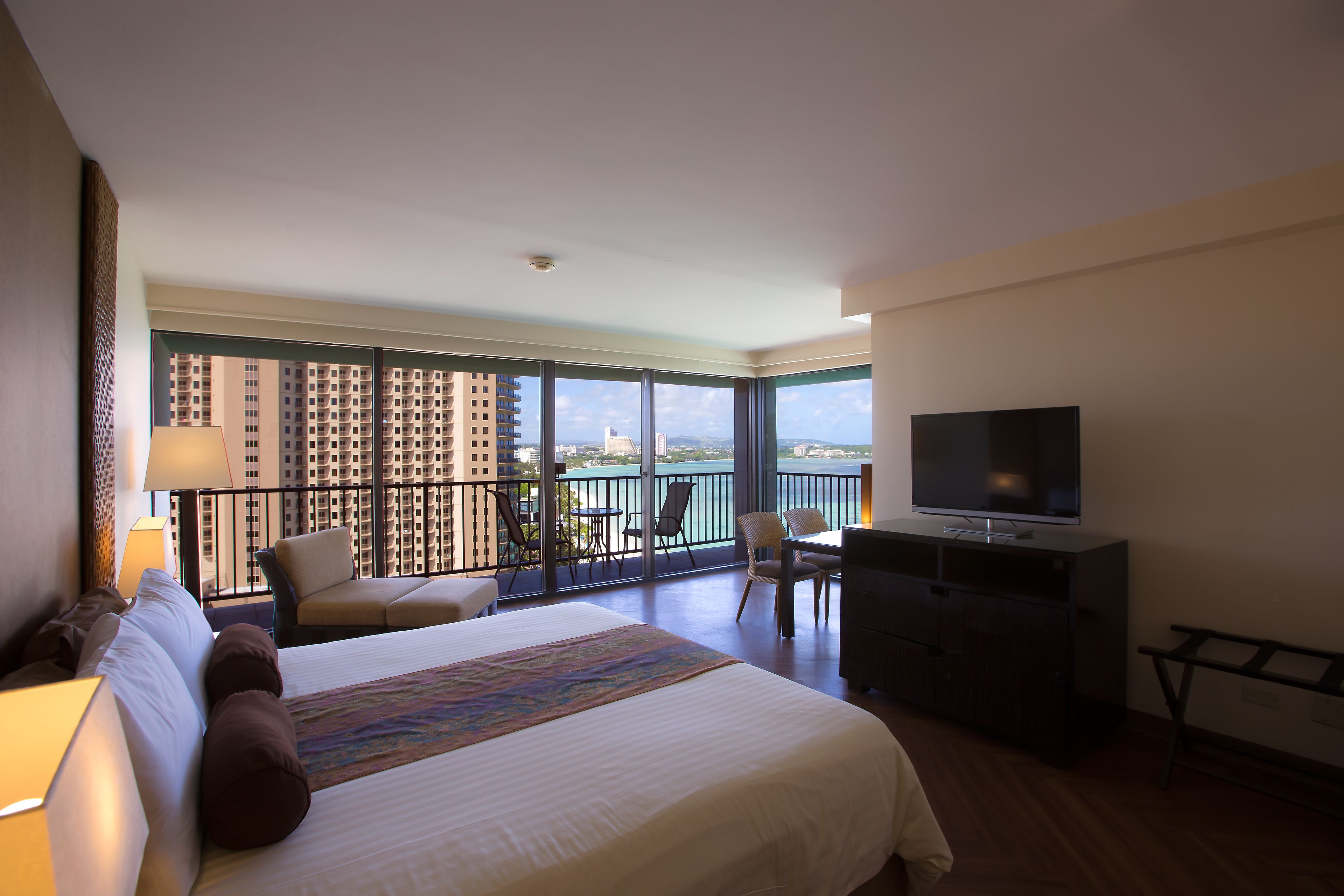 A hotel room with a large bed, a TV, a balcony with a view of the city, and a desk with a chair.