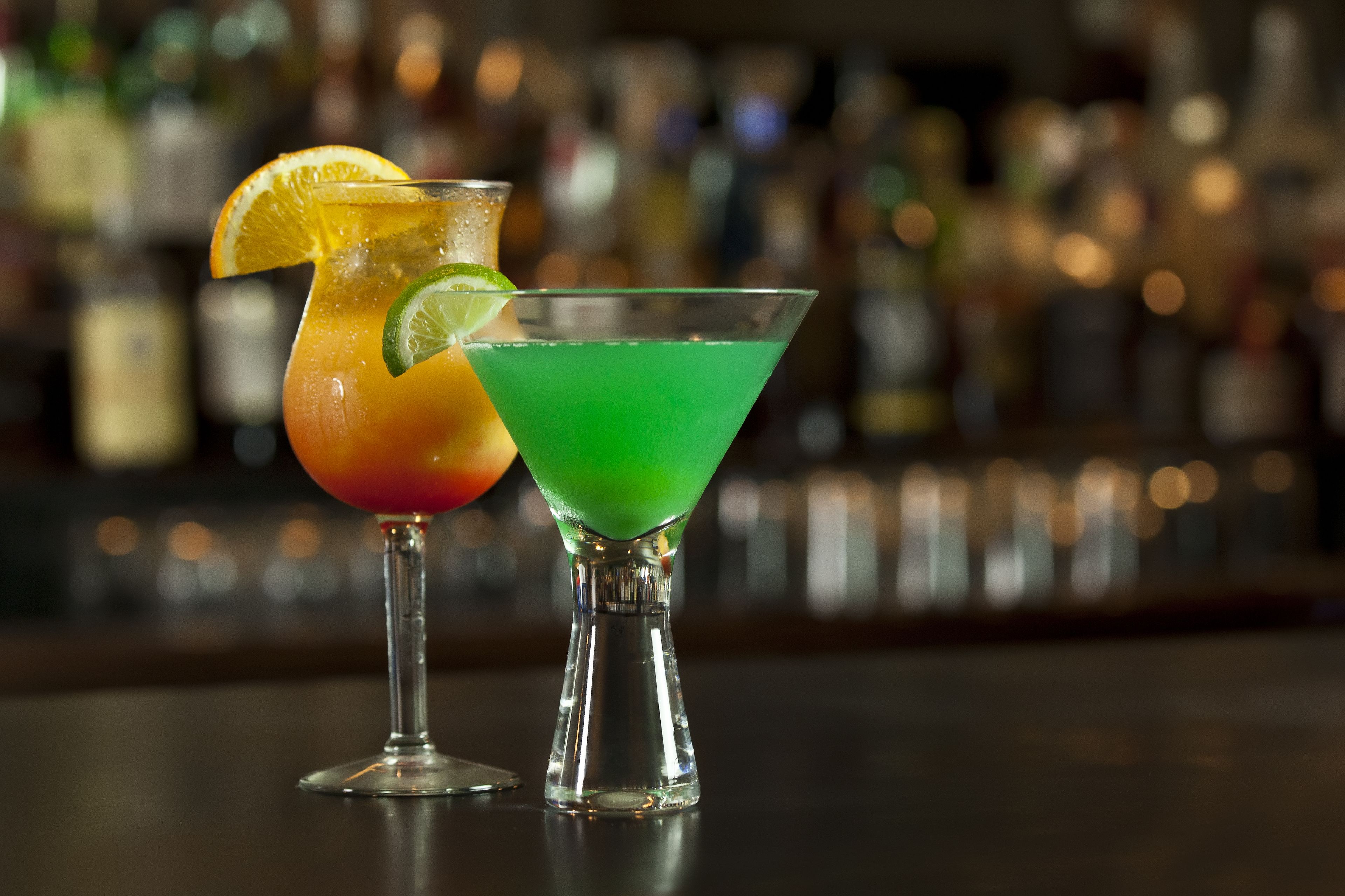 Two cocktails in martini glasses with a blurred bar background.