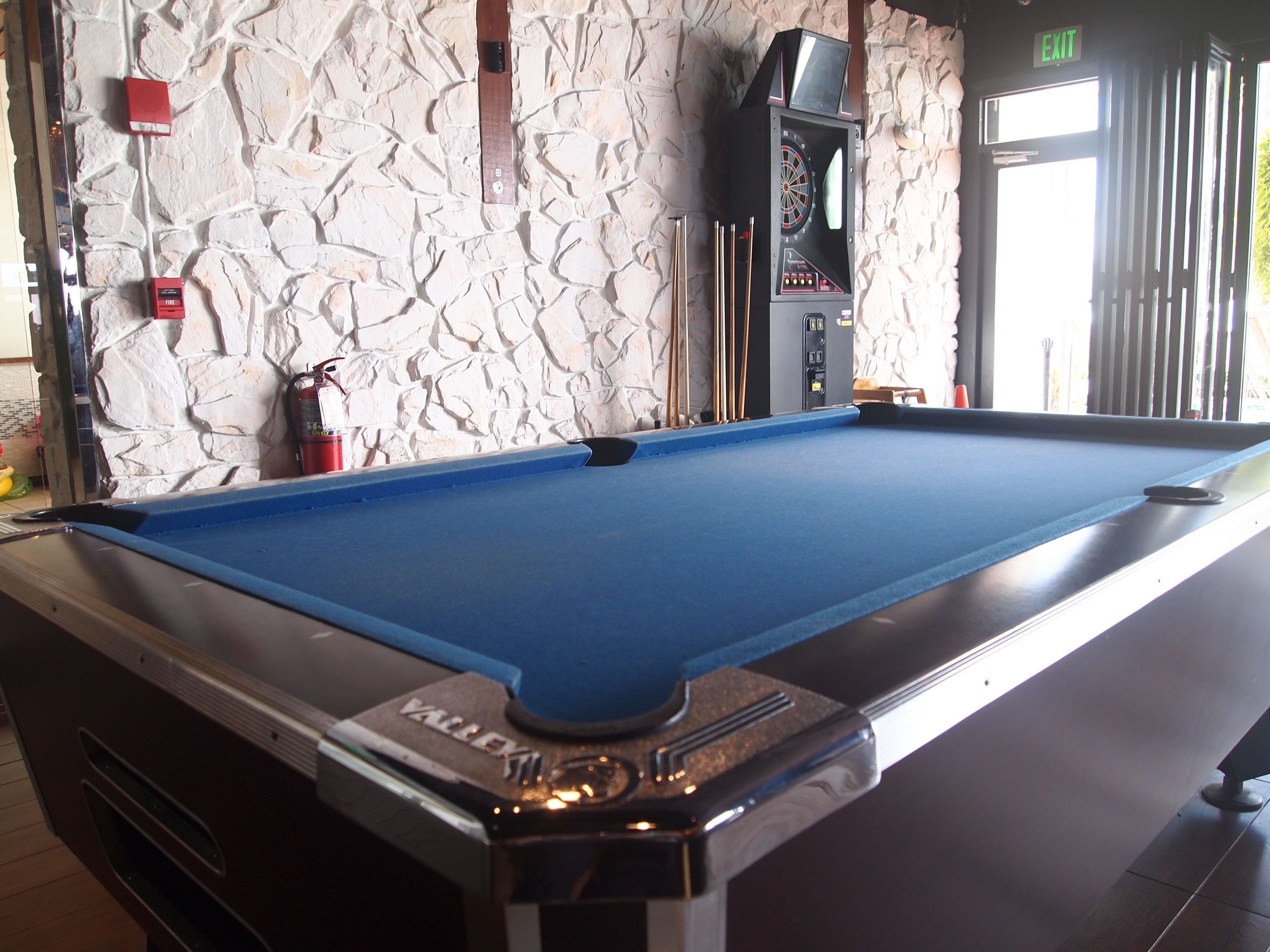 A pool table with a blue felt surface, a "Valley" logo, and a dartboard mounted above it.