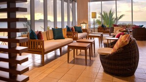 A sunlit lounge area with a sofa, chairs, and tables, overlooking a scenic view. - Guam Reef Hotel (Tamuning)
