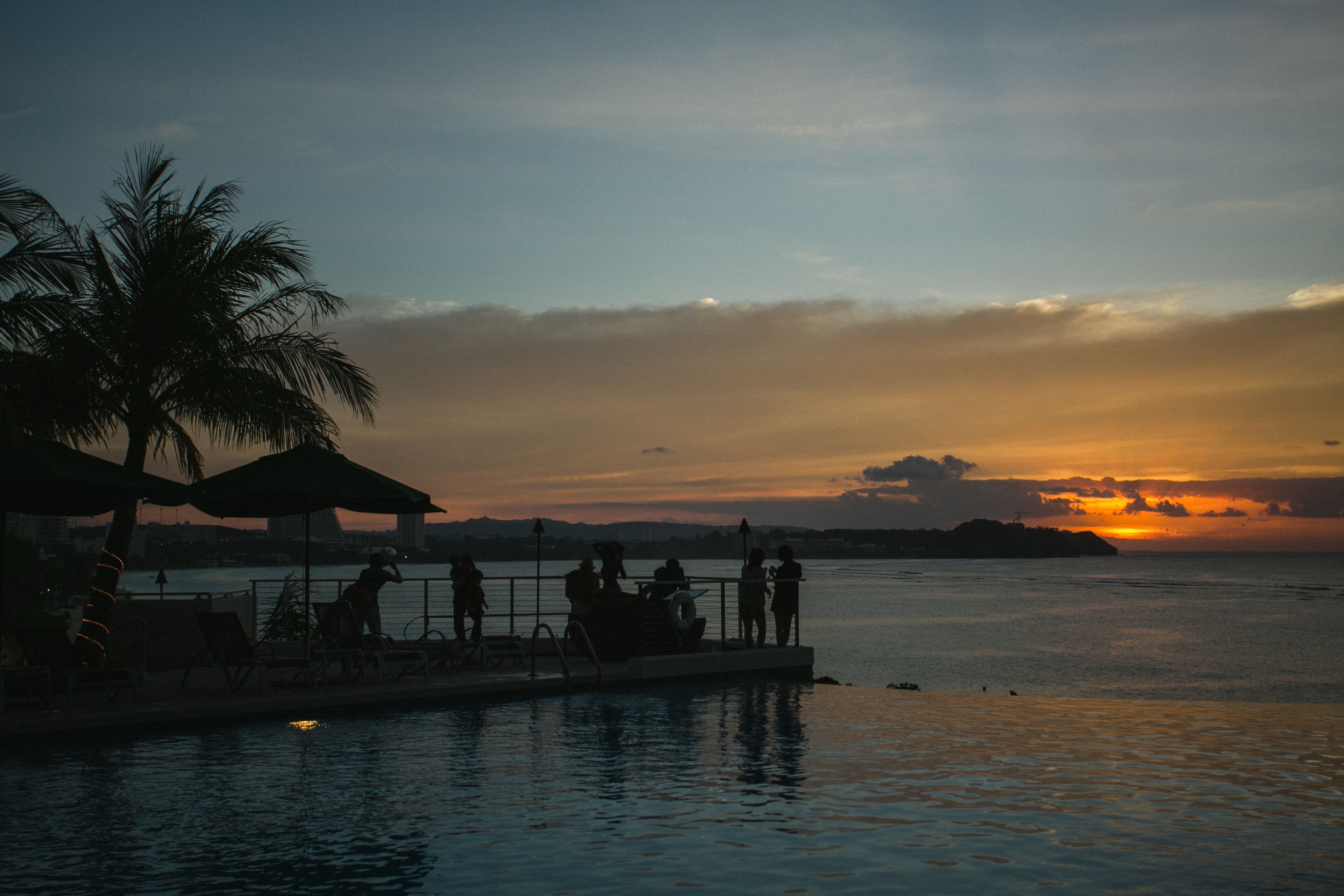 A waterfront scene with a sunset, palm trees, and people relaxing by the pool.