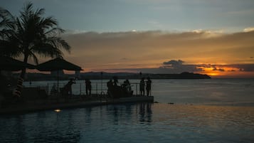 A waterfront scene with a sunset, palm trees, and people relaxing by the pool.