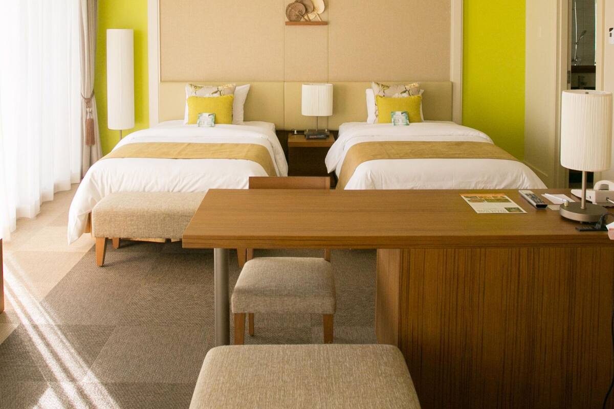 A hotel room with a TV, two beds with yellow pillows, a wooden desk, and a window.