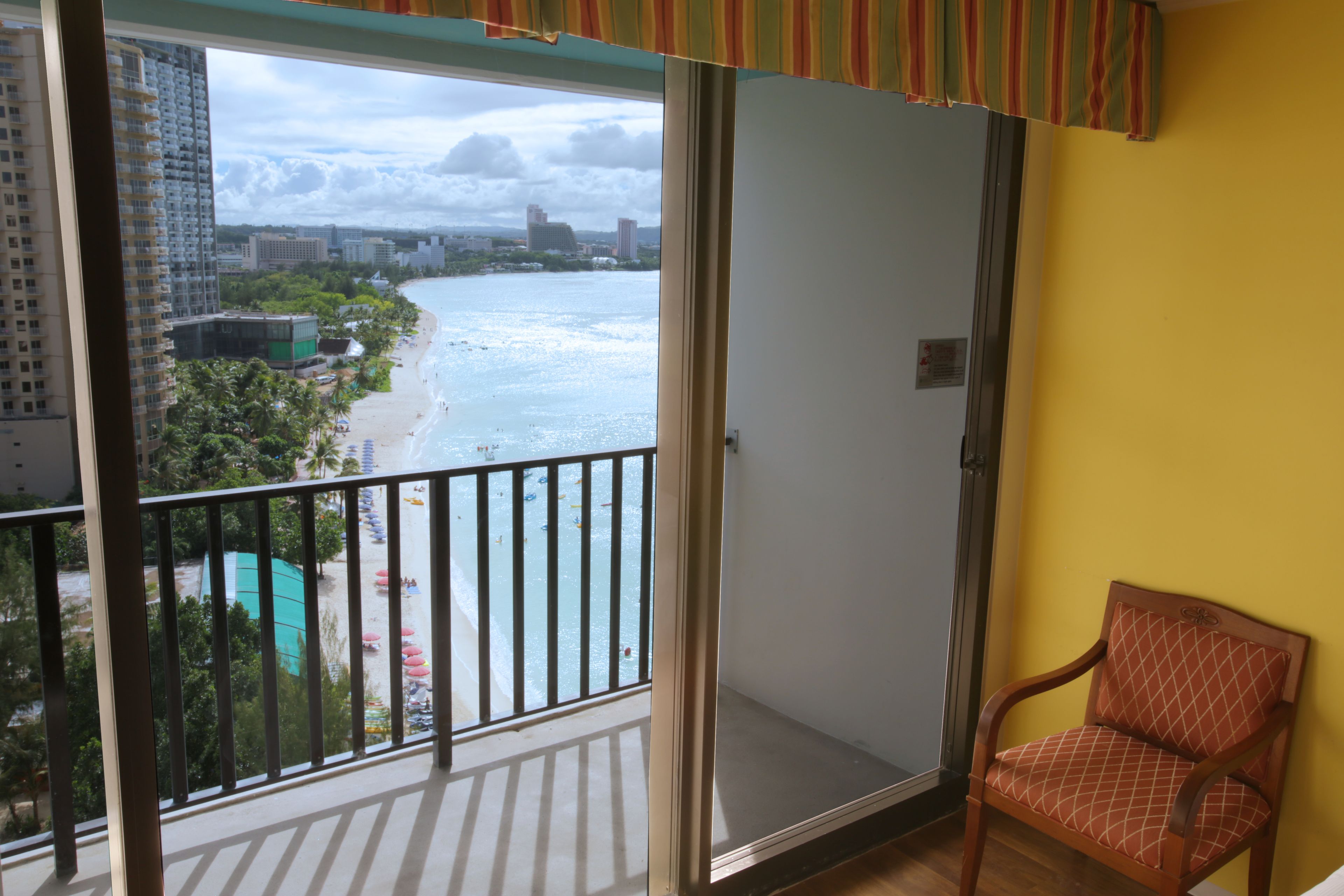 A balcony with a view of a beach and ocean, a chair, and a yellow wall.