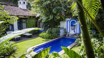 A swimming pool surrounded by lush greenery, a white building with a tiled roof, and a small gazebo.