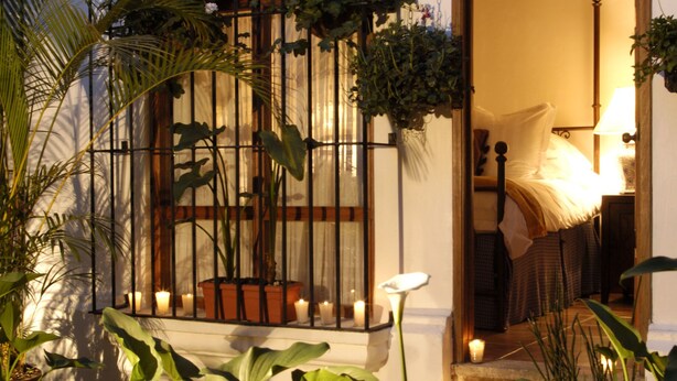 En veloplyst patio med plantekasser, et træhegn og en døråbning, der fører til et værelse med en seng og en lampe.