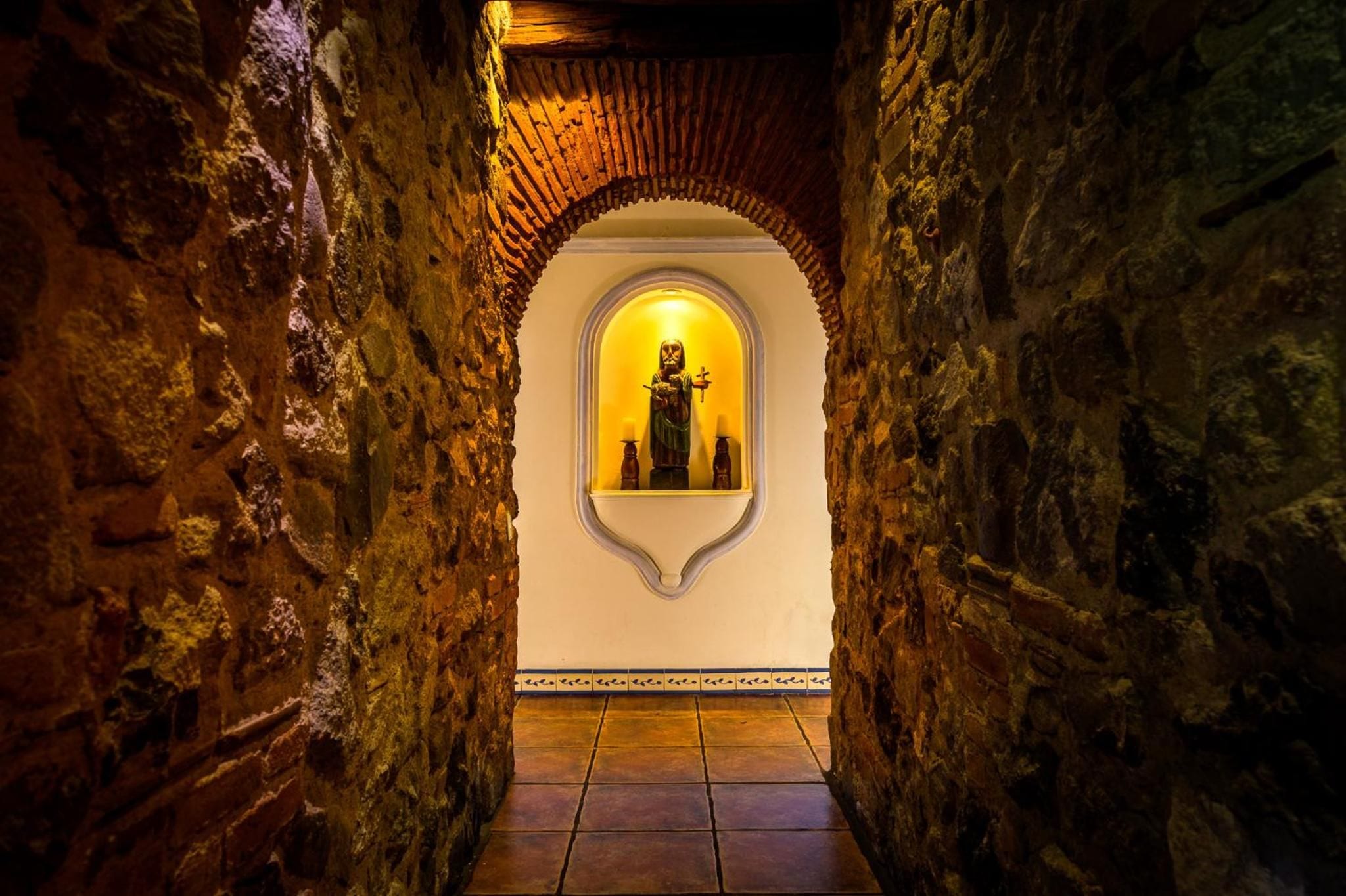 A narrow stone corridor with an arched doorway leading to a room with a statue.