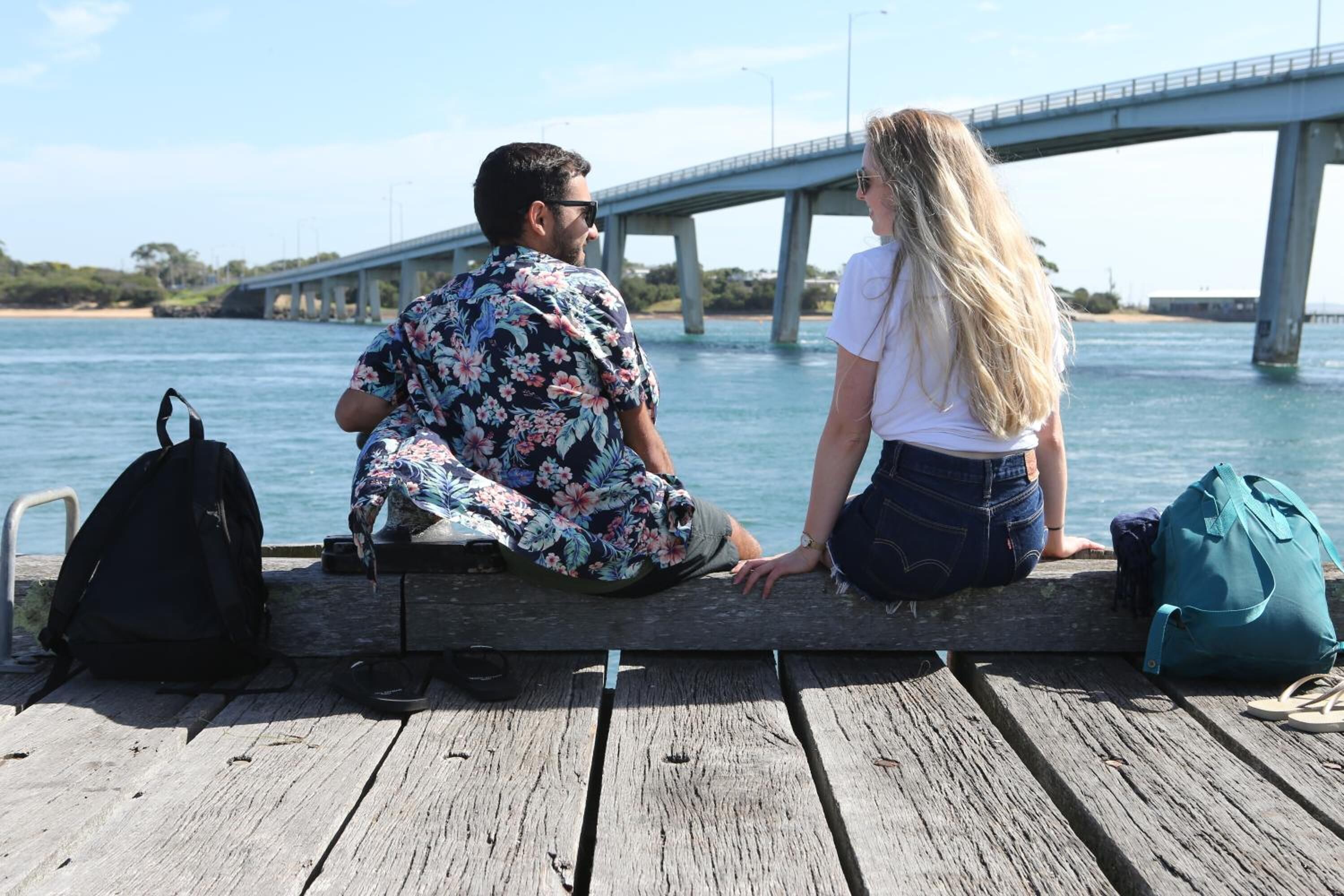 Dos personas sentadas en un muelle de madera junto a un puente sobre el agua.