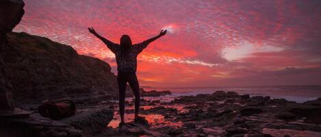 A person standing on a rocky beach with arms outstretched, facing a sunset.