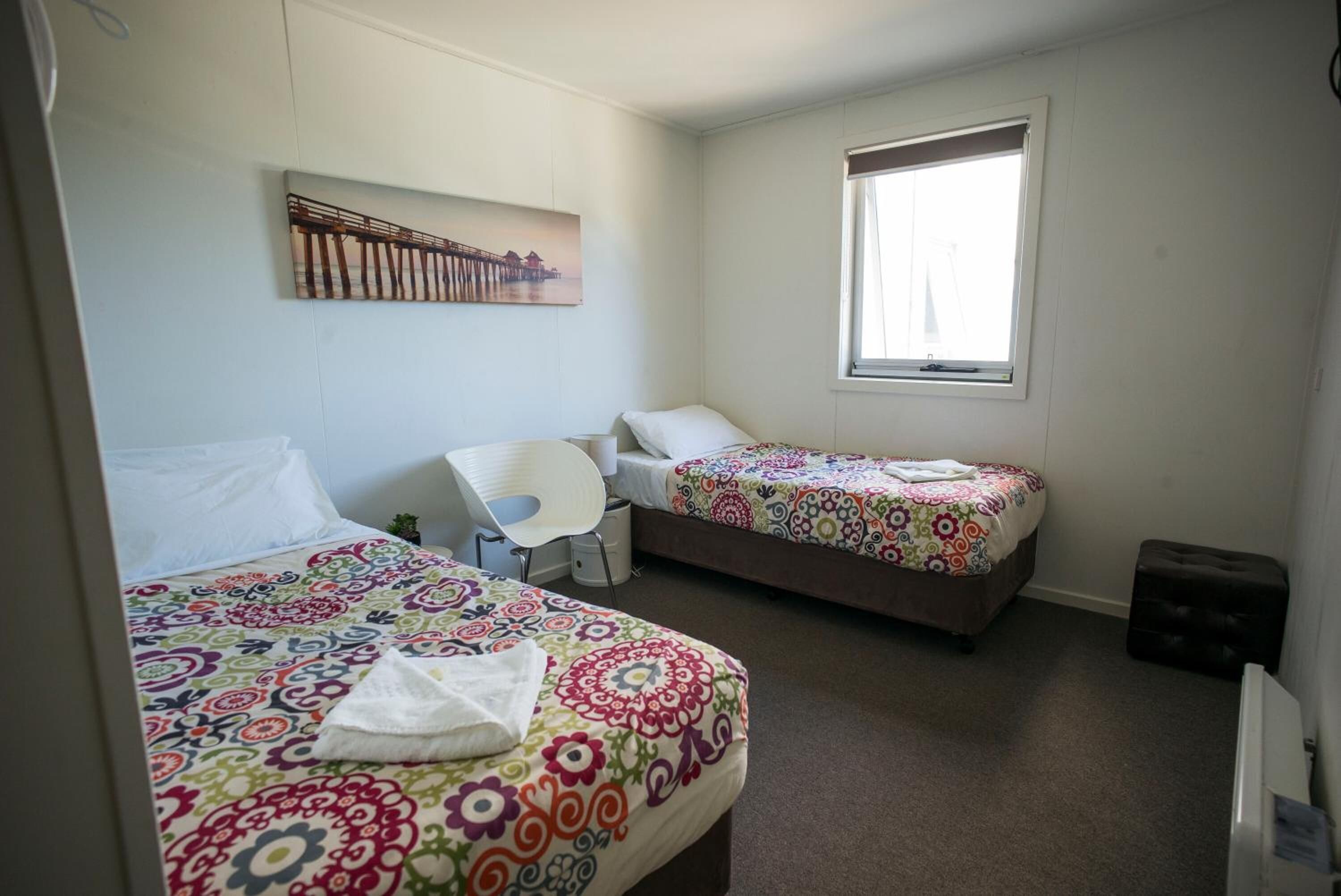 A room with two beds, one with a colorful floral bedspread and the other with a plain white bedspread. There is a chair, a small table, and a window with curtains.
