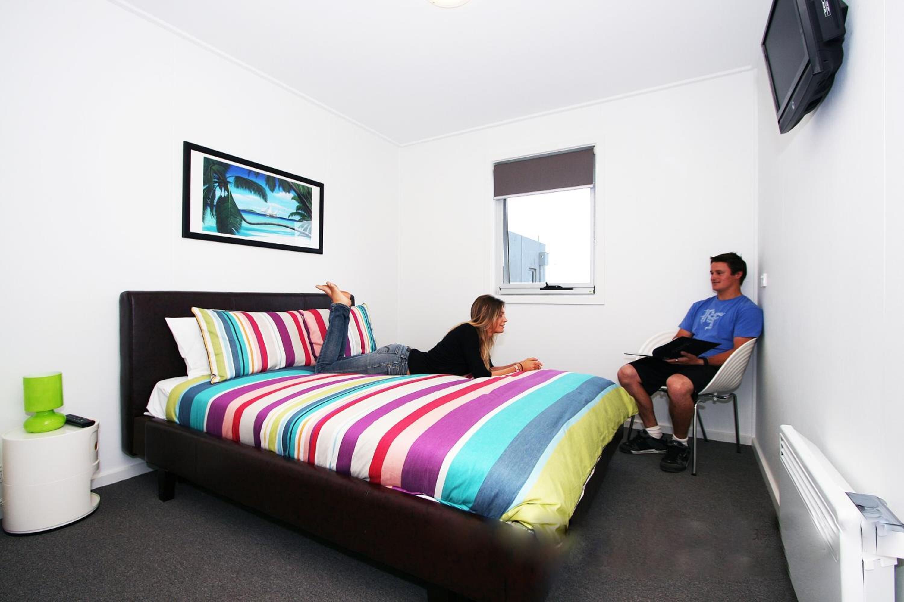 Een slaapkamer met een bed, een raam, een televisie en een klein tafeltje met een lamp.