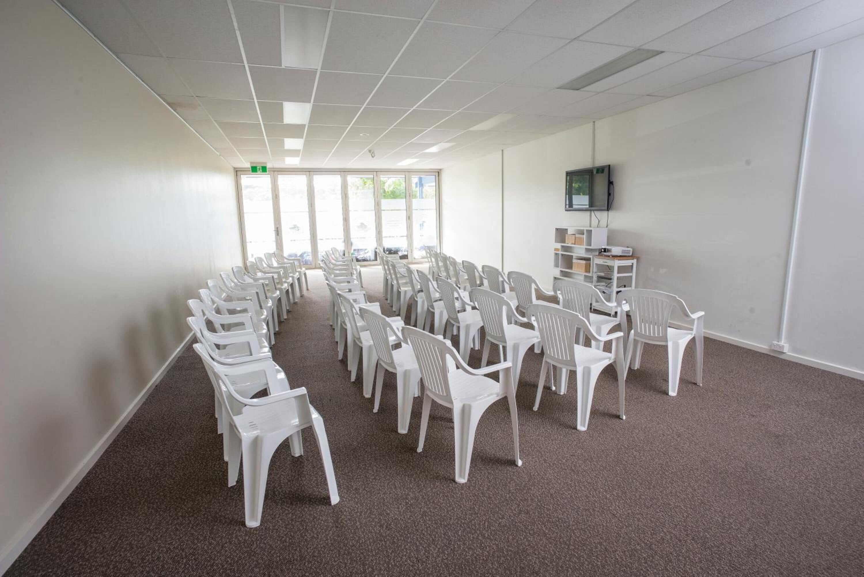 A conference room with rows of white plastic chairs, a white carpet, a white wall, a television on a stand, and a door with an exit sign.