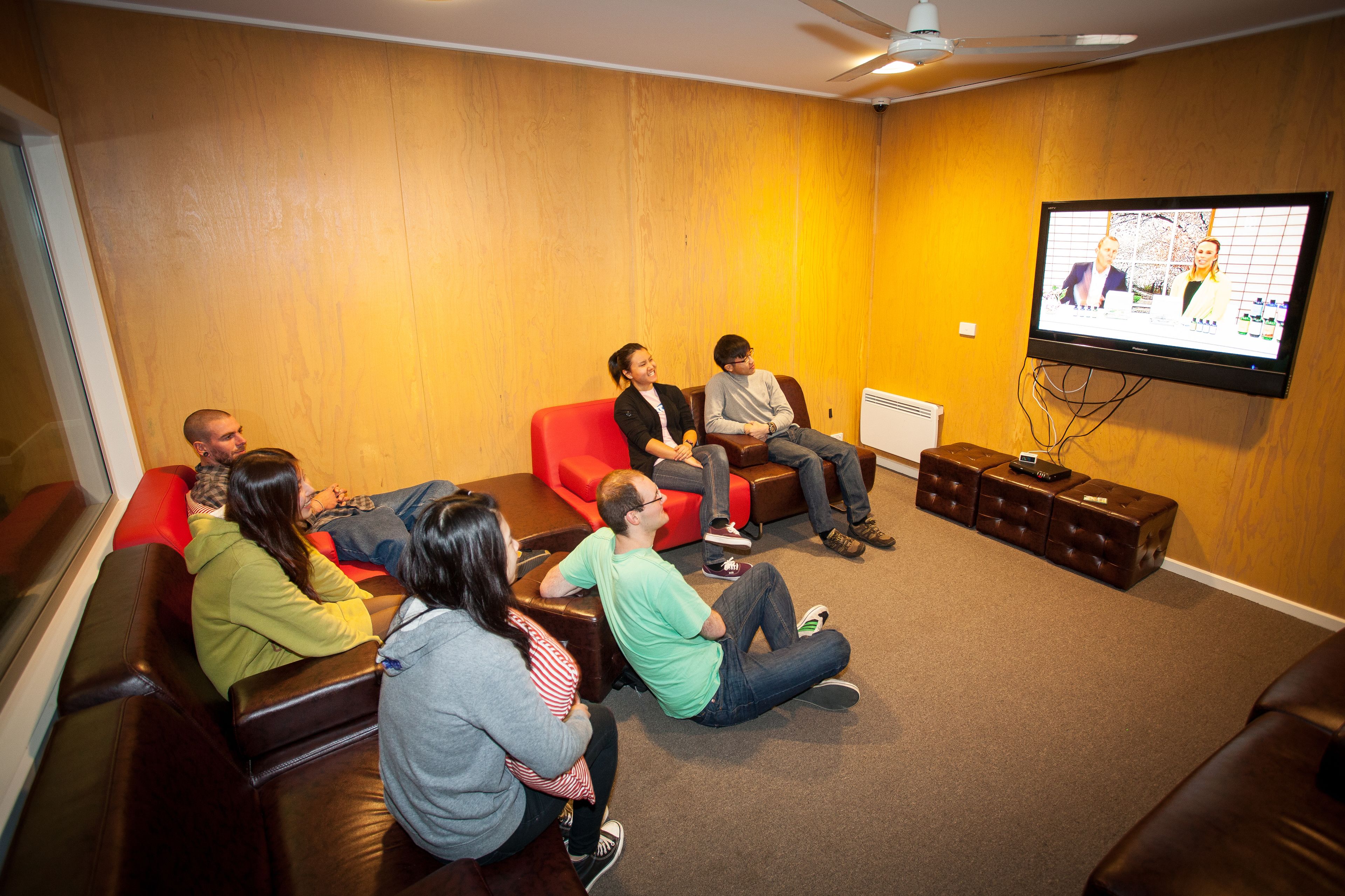 A group of people sitting on leather couches in a room with wooden walls and a flat-screen TV mounted on the wall.