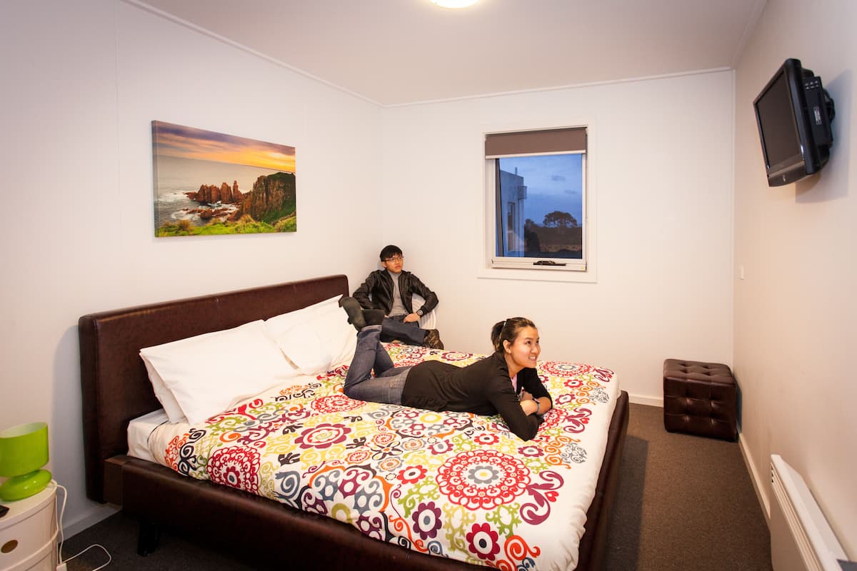 A bed with a colorful patterned bedspread, a television mounted on the wall, and two framed pictures on the wall.