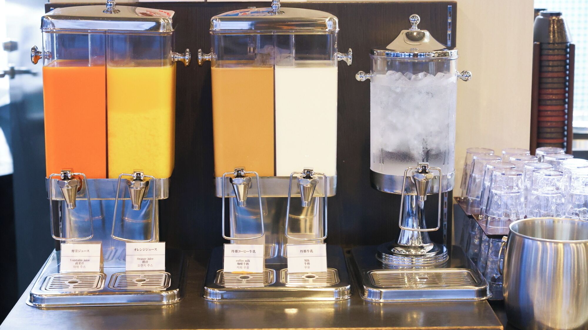 Three beverage dispensers with labels for vegetable juice, orange juice, and coffee milk.