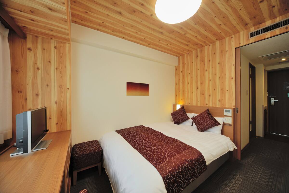 A hotel room with a wooden ceiling, a bed with a patterned bedspread, a bedside table with a flat-screen TV, and a small bench.