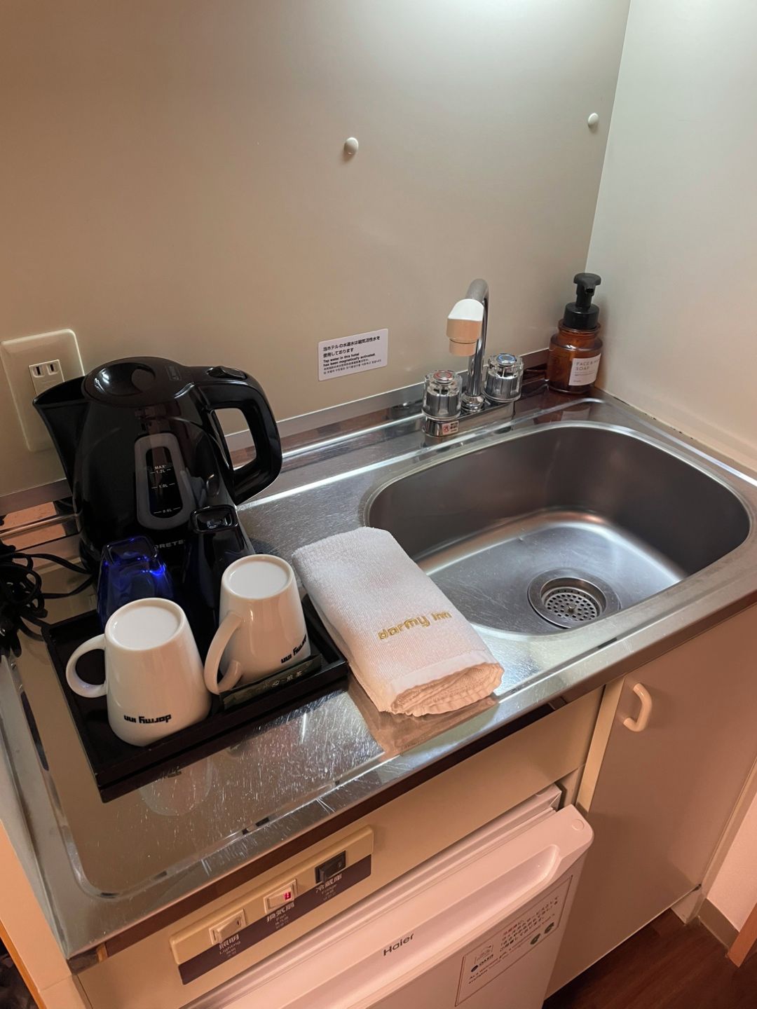 Eine Hotelküche mit einer Kaffeemaschine, zwei Tassen, einem Handtuch und einer Seifenspender.