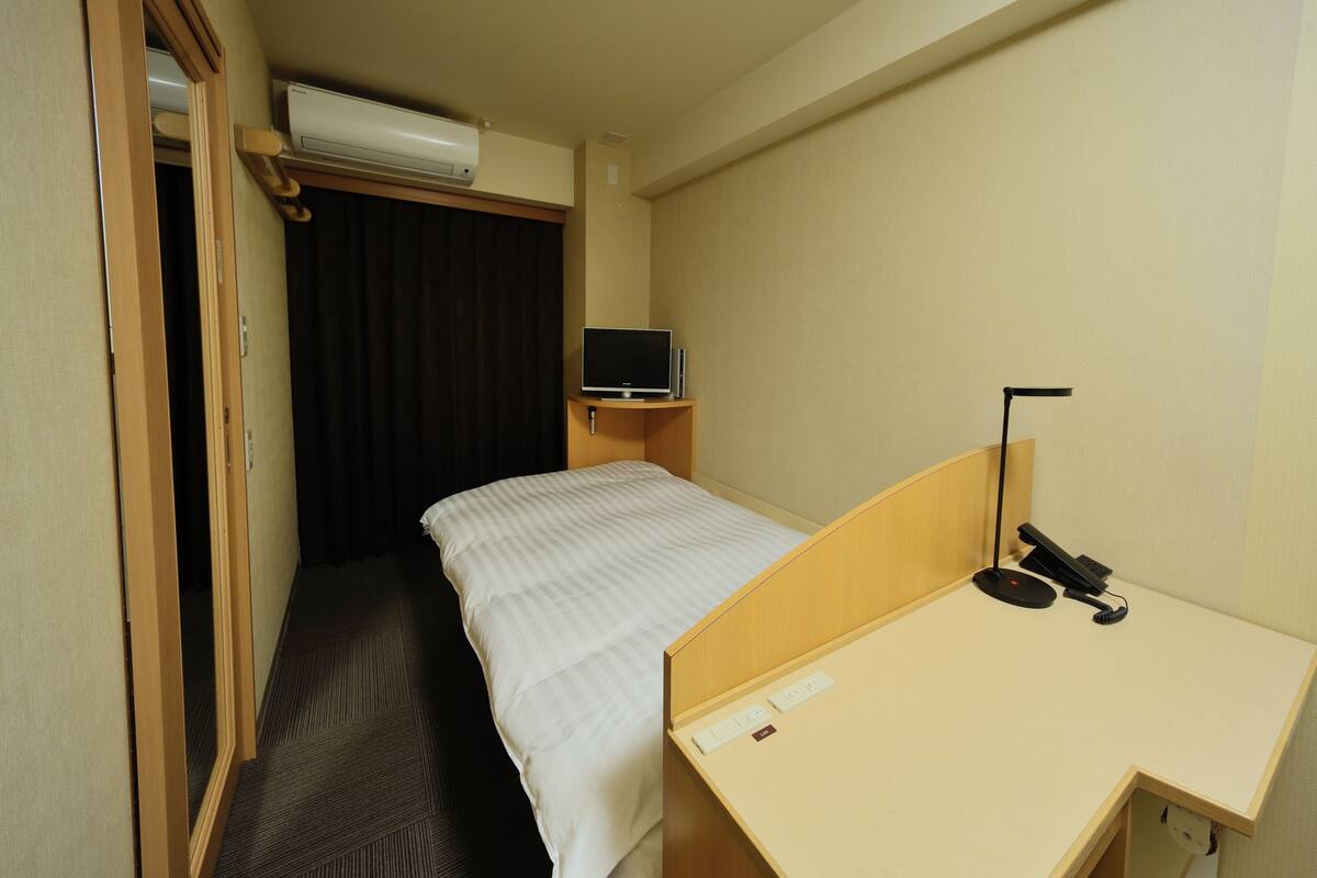 A hotel room with a single bed, a desk with a computer, and an air conditioning unit above the bed.