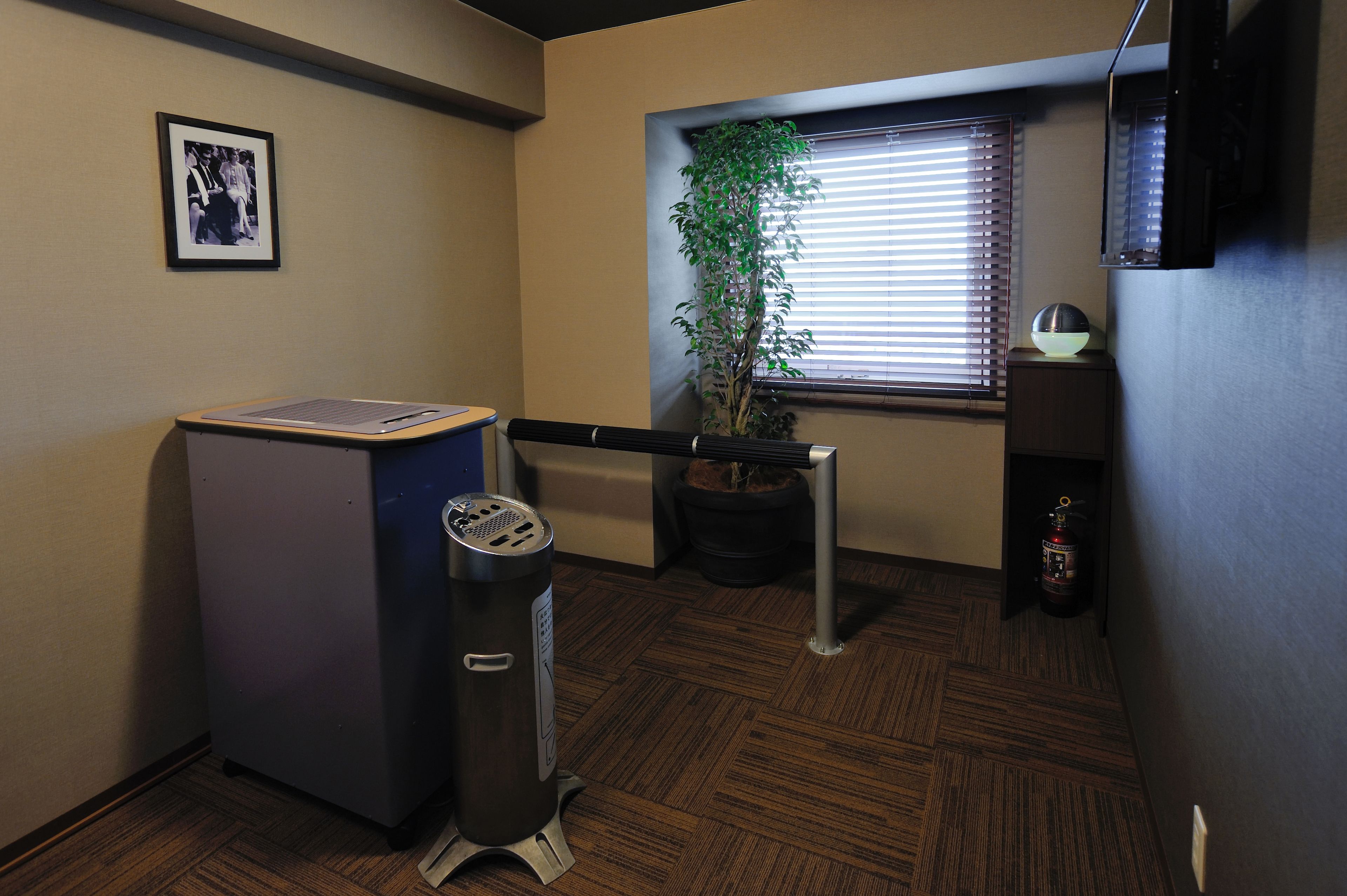 A small office with a filing cabinet, a trash can, a potted plant, a window with blinds, and a fire extinguisher.