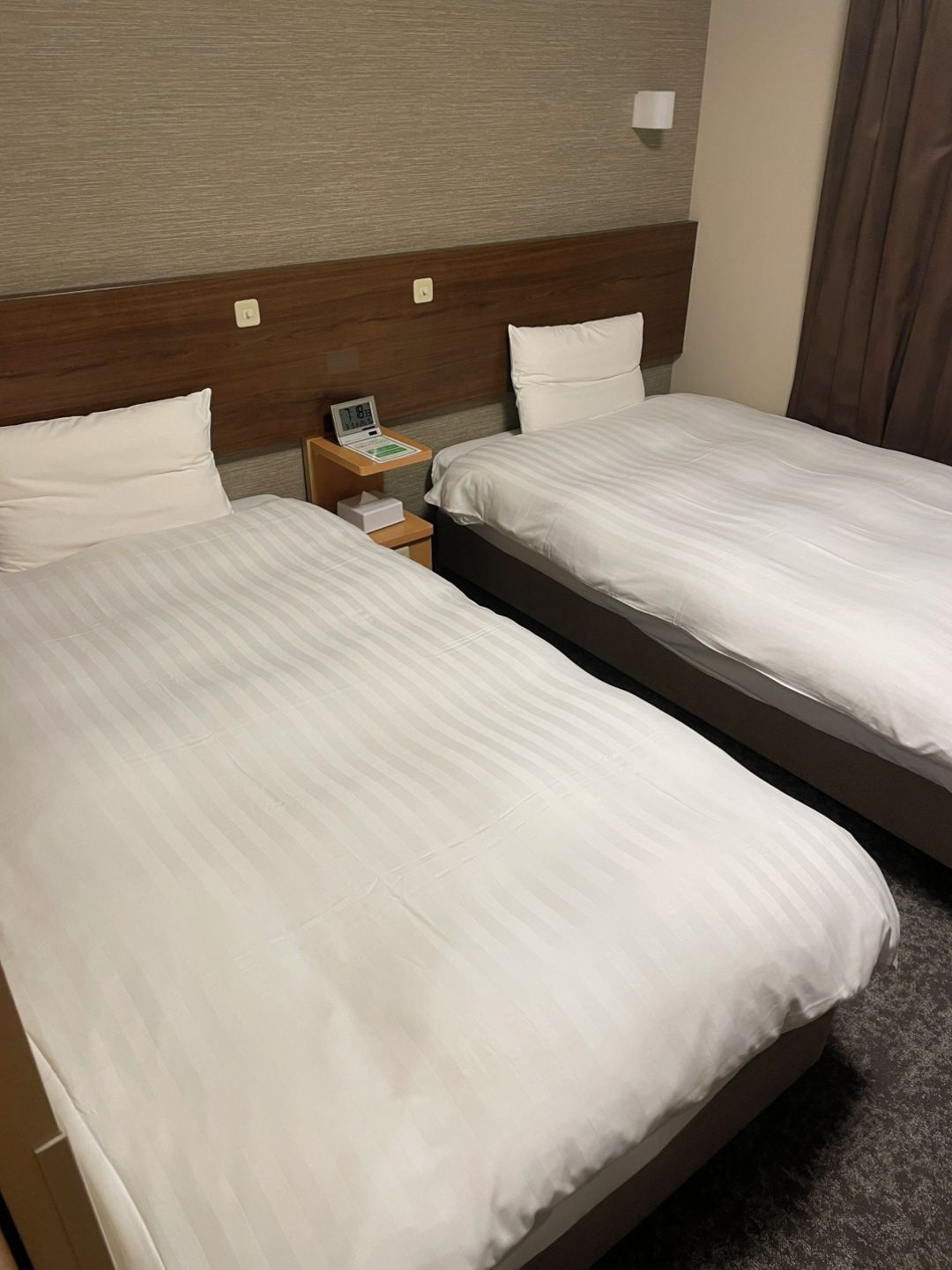 Quarto de hotel com duas camas de solteiro, mesa de cabeceira com relógio e cabeceira de madeira.