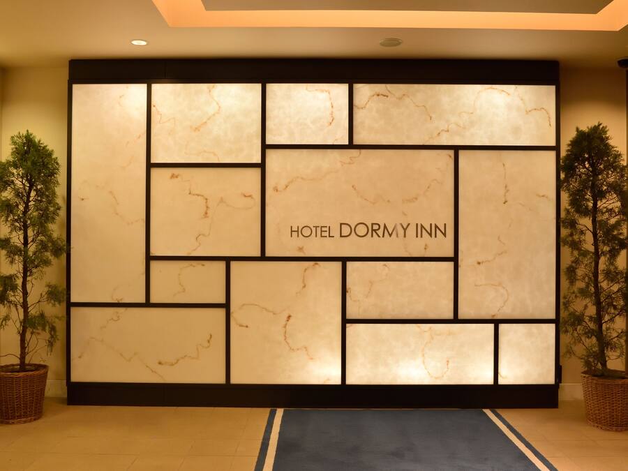 A reception area with a marble wall and the text "HOTEL DORMY INN" in the center. There are two potted trees on either side of the wall.