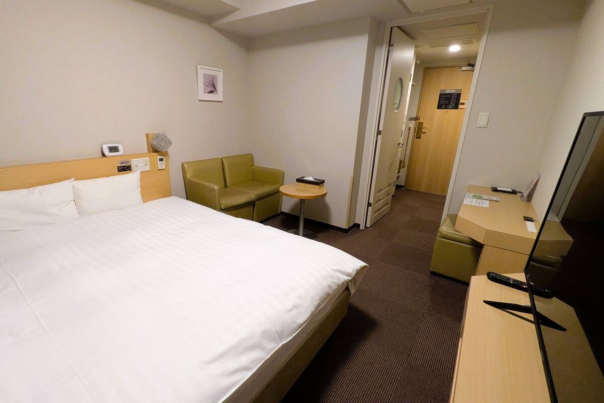 A hotel room with a bed, a desk, a chair, a small table, a TV, and a door leading to another room.